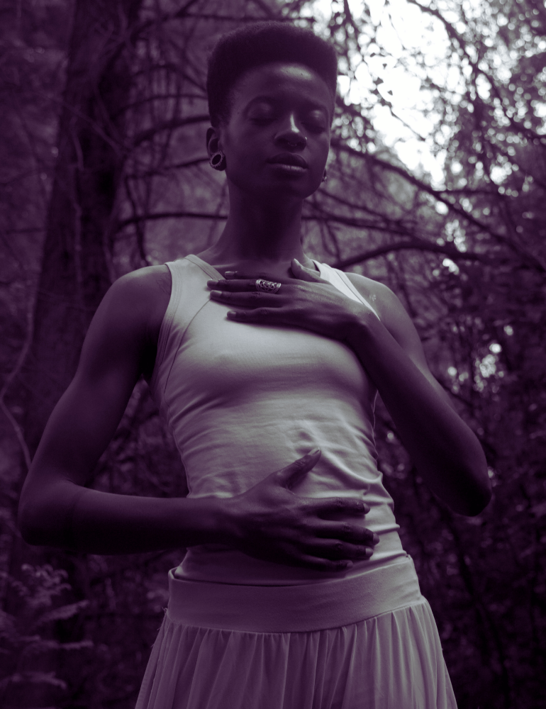 Person in white casual clothes standing in a forest with their hand on their womb and chest