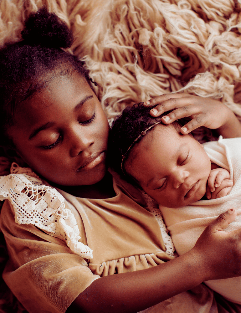 Young girl lying on a fluffy beige rug holding a baby wrapped in a blanket.