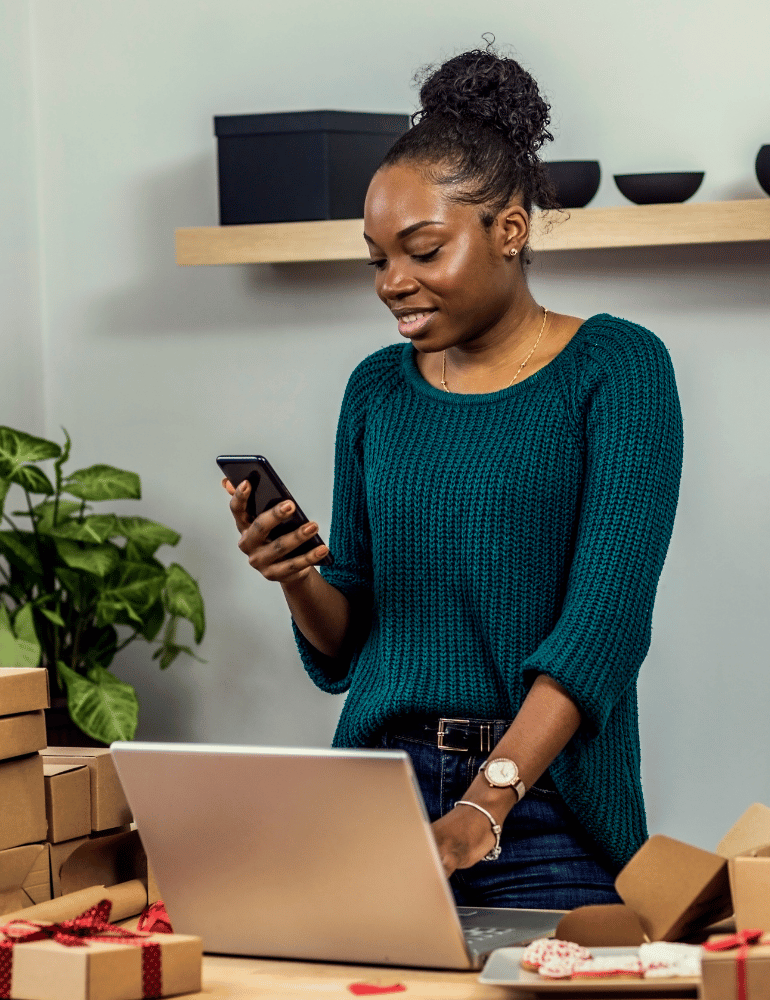 Woman using a smartphone in front of a laptop with boxes and a plant in the background