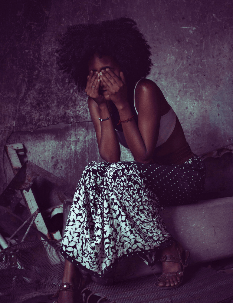 Woman sitting against a textured wall with a patterned skirt with her hands over her eyes in distress