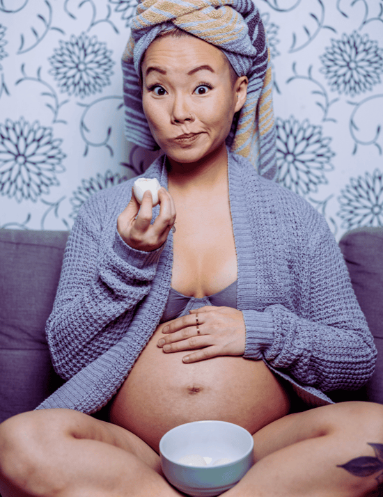 Pregnant woman sitting on a couch with a towel on her head, holding a bowl while eating an egg.
