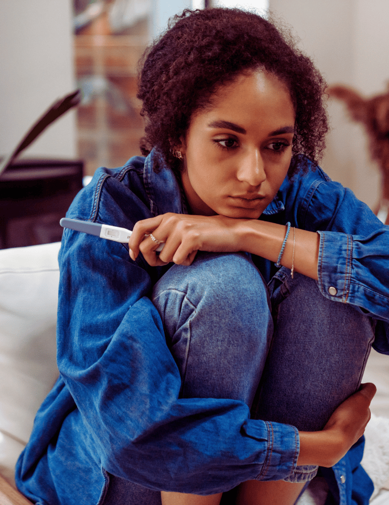 Person sitting on a couch holding a purple cushion, wearing a blue denim jacket holding a pregnancy test.