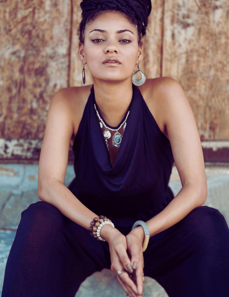 Woman in a dark blue linen jumper with jewelry sitting against a textured wall.