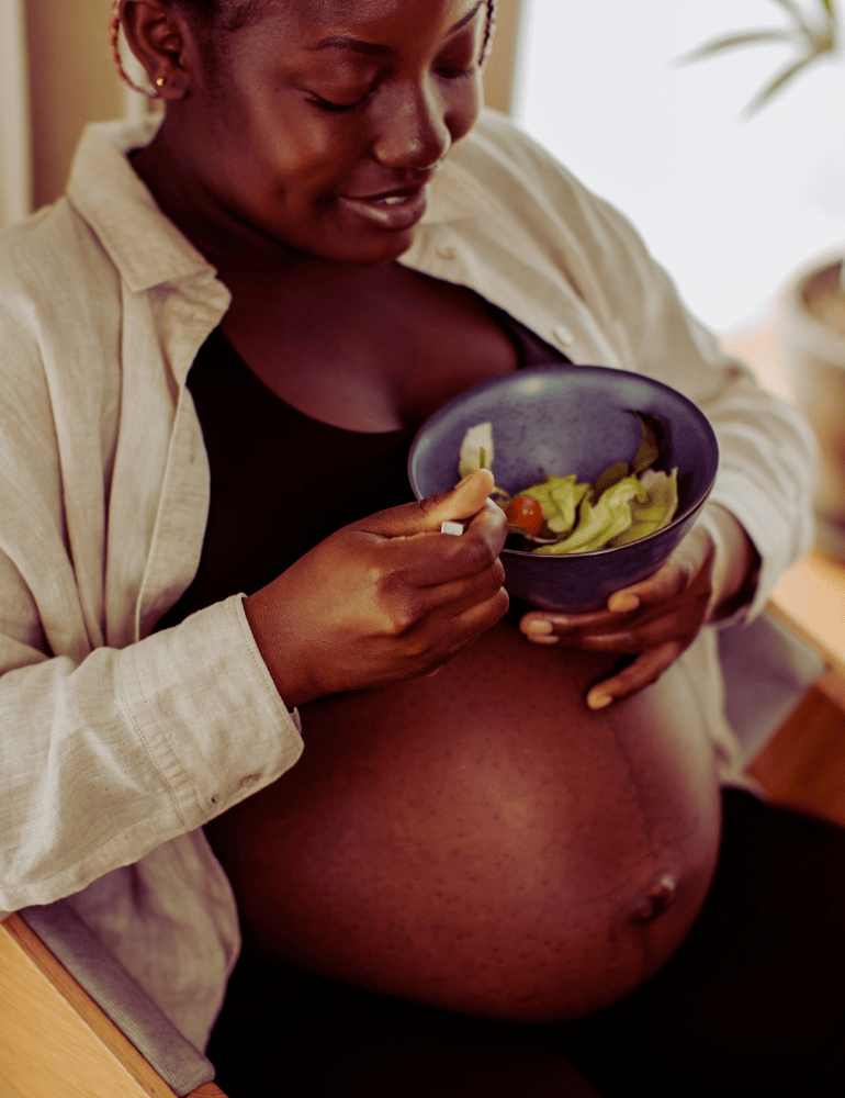 Pregnant woman eating a salad from a purple bowl indoors.