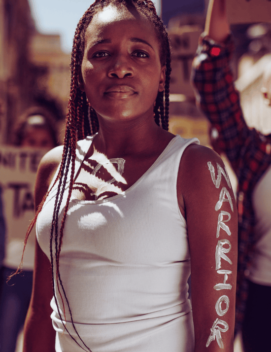 Woman with braided hair wearing a white sleeveless top with 'WARRIOR' written on her arm, standing in an outdoor setting.