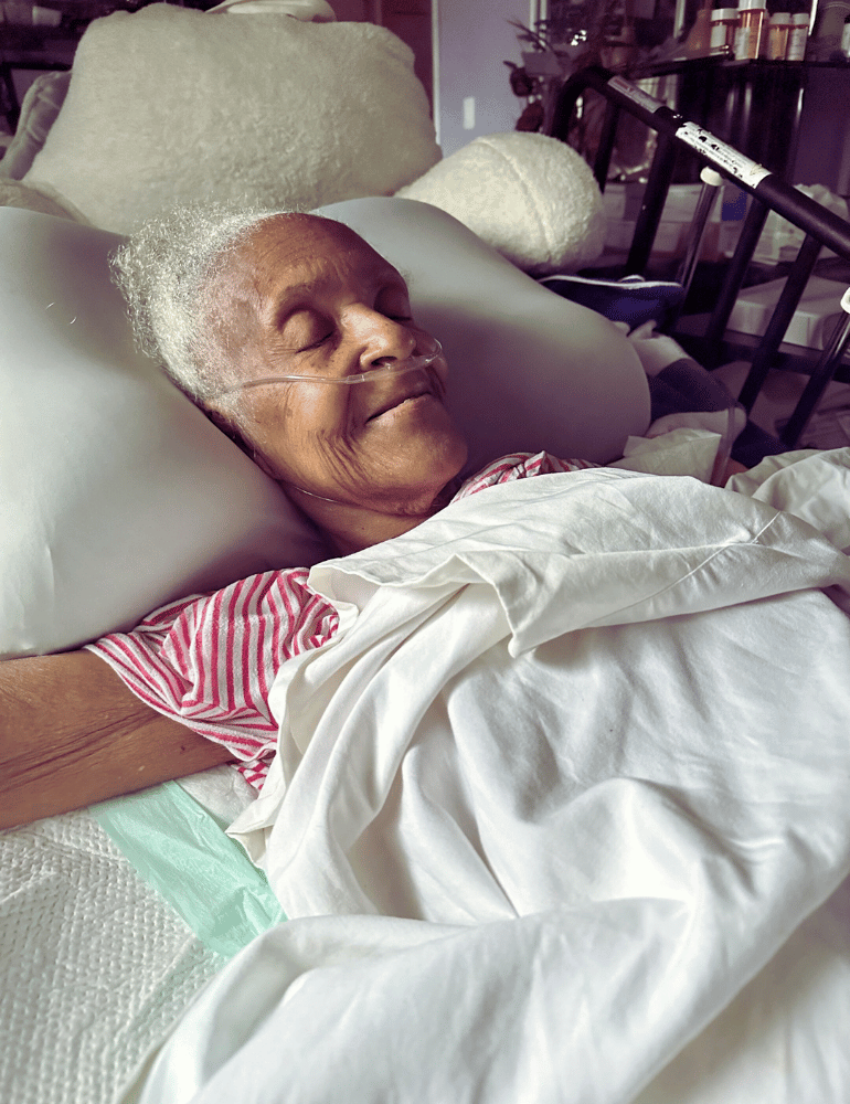 Person lying in bed with white pillows and a blanket, wearing a pink and white striped shirt.