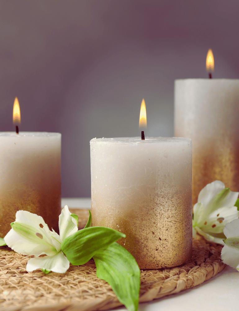 Three white candles with a gradient effect on a woven mat with white flowers.